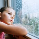 Little Sad Girl Pensive Looking Through The Window Glass With A Lot Of Raindrops. Sadness Childhood